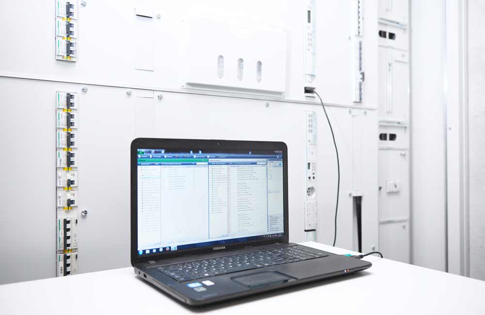 Ordinateur connecté à un grand tableau électrique dans un environnement de travail, montrant l'intégration technique du bâtiment par Flückiger.